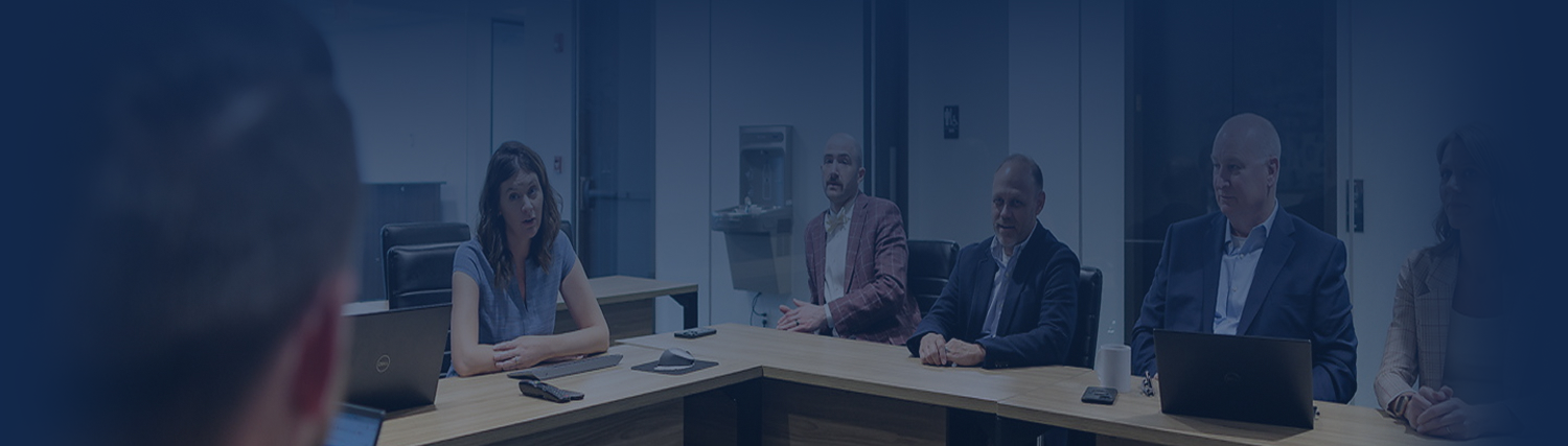 Five people in business attire sit around a conference table in a modern office, listening to a speaker out of frame; some have laptops open on the Component Page, and the room features glass walls.