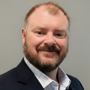 Aaron Winters, a man with short light brown hair and a beard, smiles at the camera in a navy blazer over a white checkered dress shirt, set against a plain gray background.