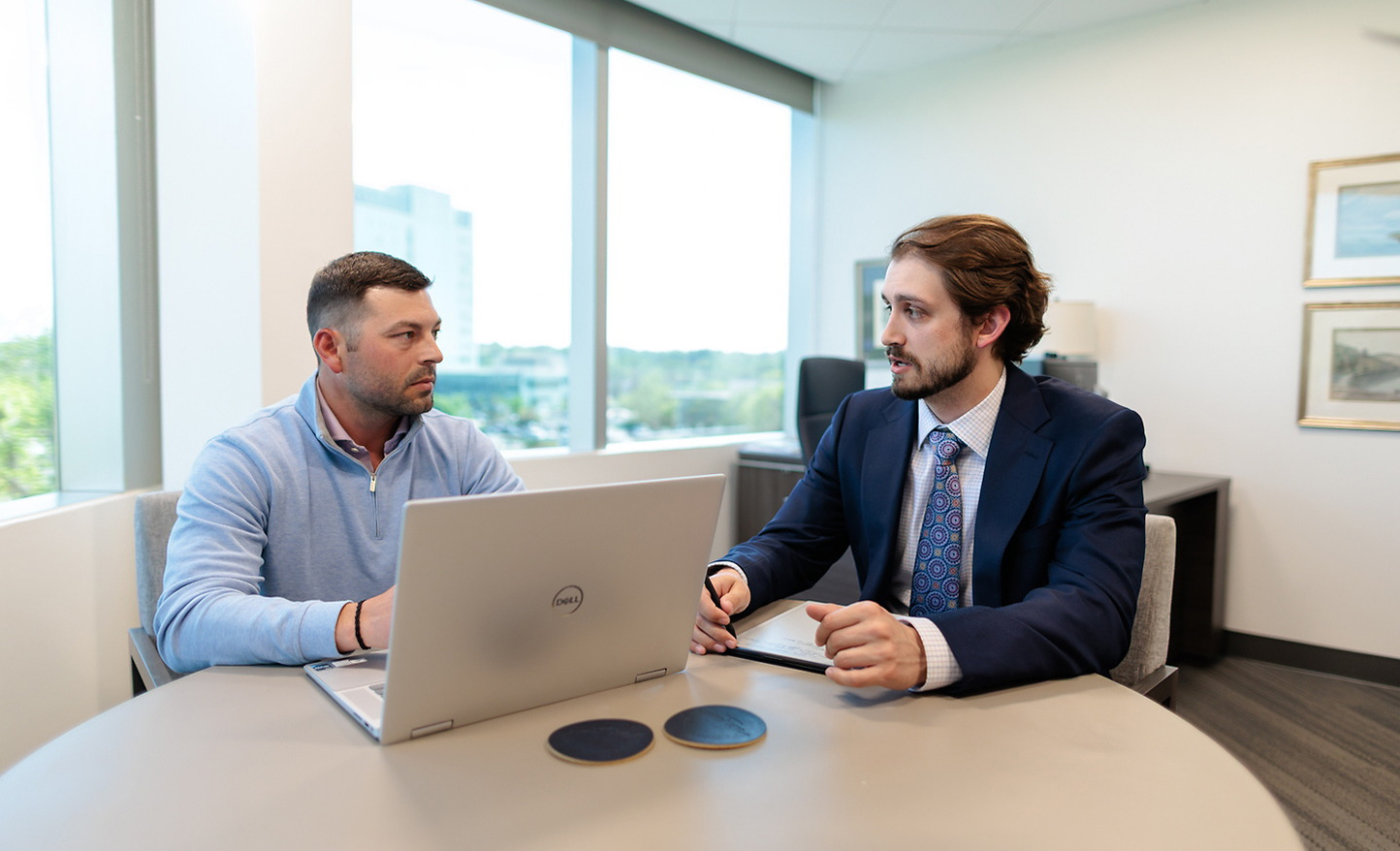 Two men sit at a round table in a modern office. One, a financial advisor from Omaha in a suit and tie, gestures as he discusses plans with his client, who faces a laptop. Large windows and framed art are visible in the background.