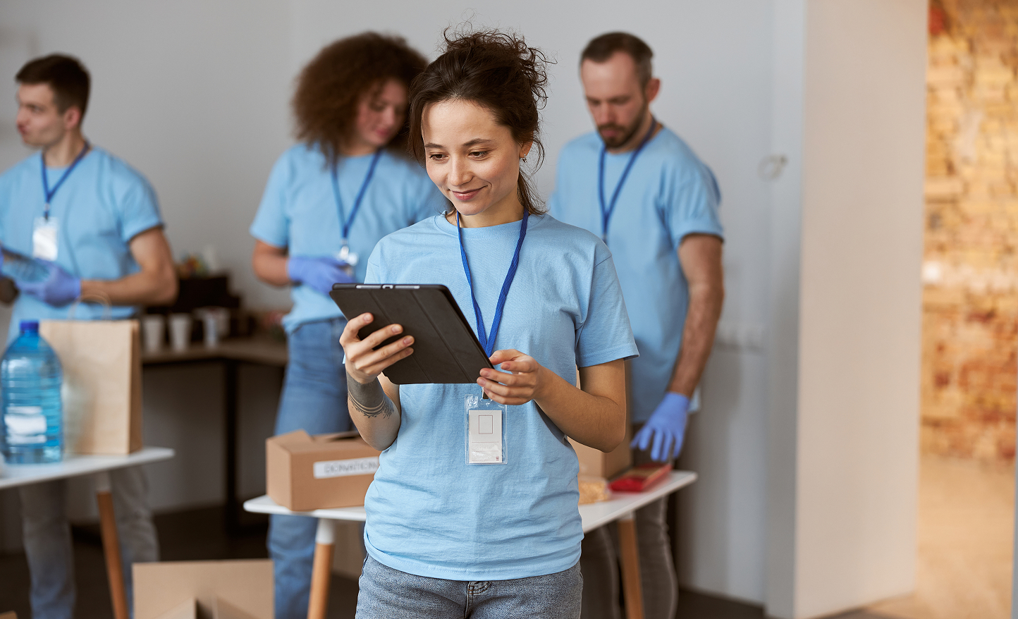 A young woman in a blue t-shirt and name badge smiles while holding a tablet. Nearby, a team in matching shirts organizes supplies and boxes, reflecting the teamwork found at a financial advisor Omaha office during volunteer activities.