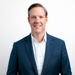 A man with short, light brown hair wearing a blue plaid suit jacket over a white dress shirt stands smiling confidently against a plain white background.