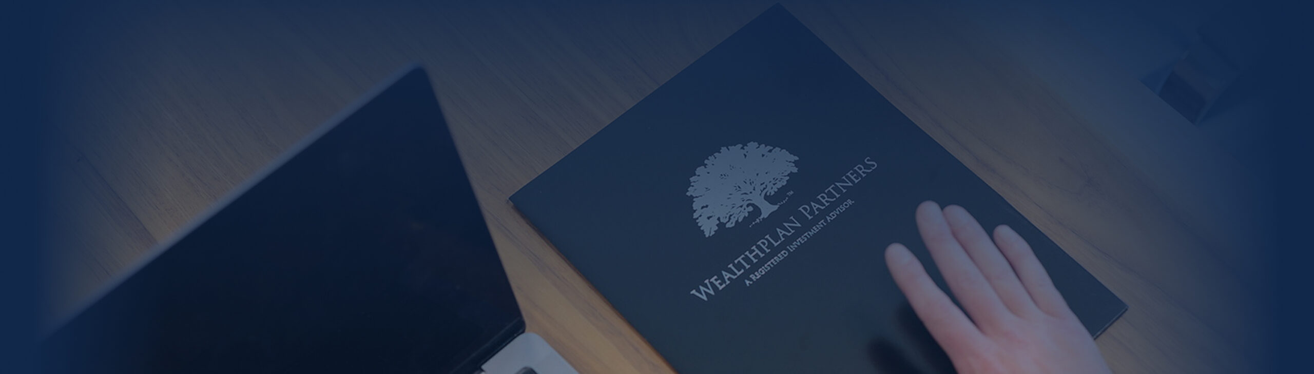 A hand rests on a dark folder labeled WealthPlan Partners with a tree logo, beside a partially visible laptop on a wooden desk, suggesting financial planning.
