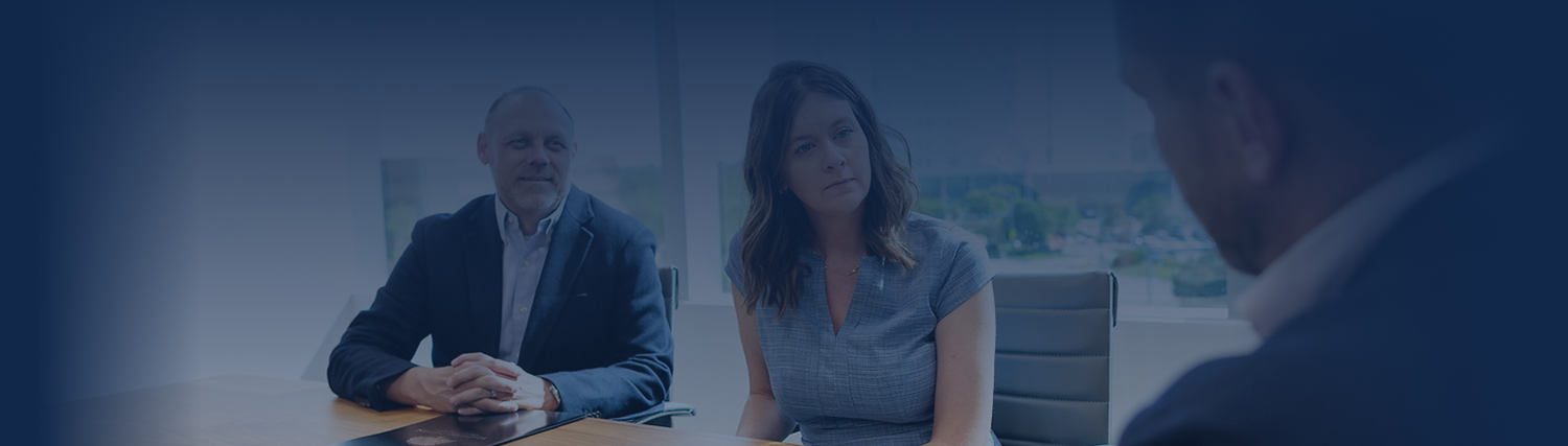 Two people sit at a conference table, attentively listening to a third person whose back is to the camera. The setting appears to be a modern office with large windows, suggesting an estate planning meeting in progress.