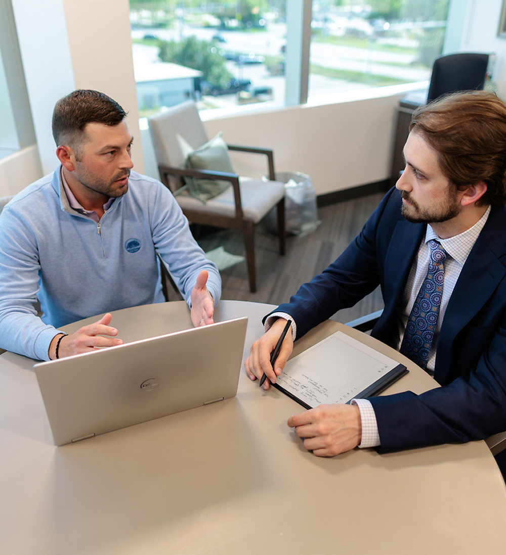 Two men sit at a round table in an office, discussing estate planning. One man in a blue sweater gestures while speaking, while the other in a suit listens with a notepad. A laptop and papers are on the table, with large windows in the background.