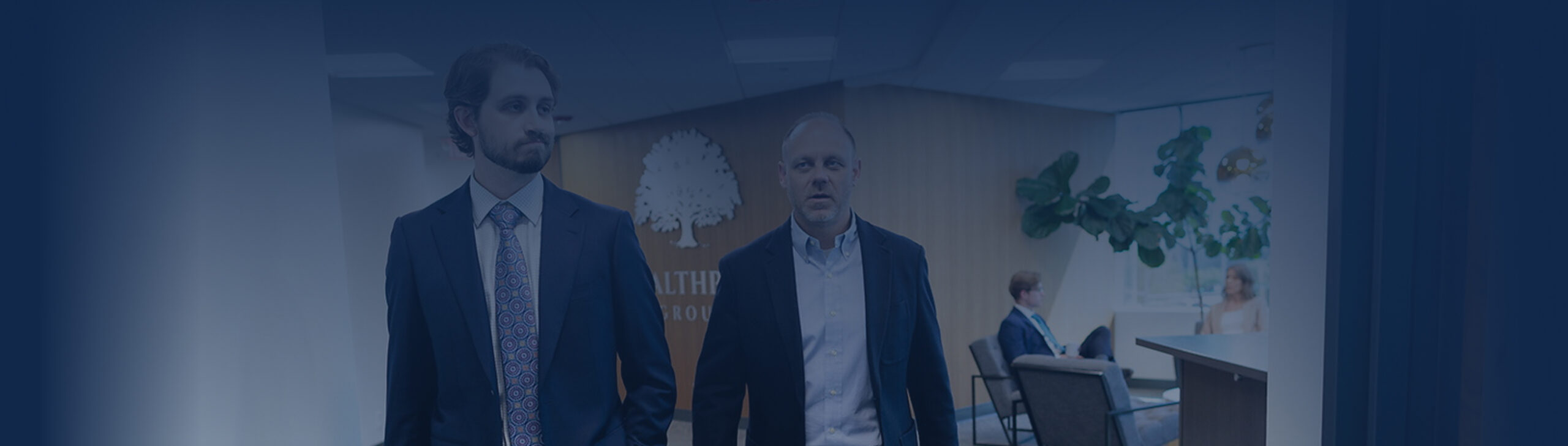 Two men in business attire walk through an office lobby featuring a tree logo on the wall behind them. In the background, two people are seated by a reception desk with indoor plants and a "Find An Advisor" sign visible.