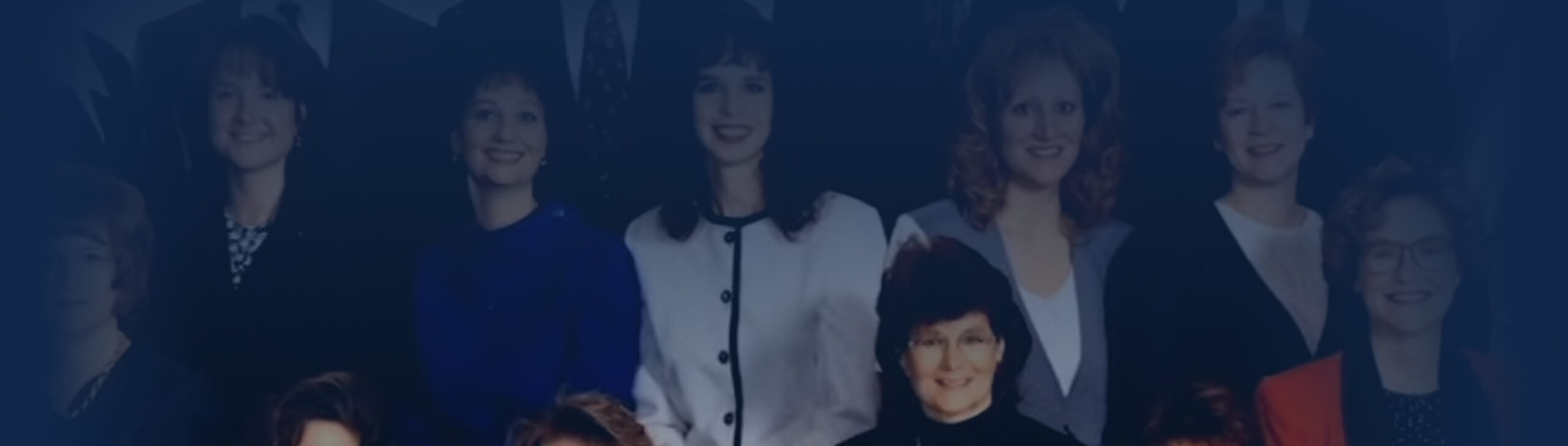 A group of women dressed in formal and business attire pose for a group photo, smiling at the camera against a dark background, reflecting the firm history.