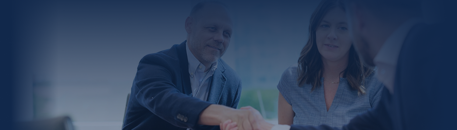 A man and woman sit at a table, smiling and shaking hands with another person in a professional setting, suggesting a business meeting or agreement about insurance solutions.