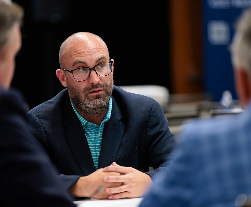 A man with glasses and a trimmed beard, wearing a navy blazer and striped shirt, sits at a table with hands clasped, engaged in an insurance planning discussion with two others in a meeting setting.