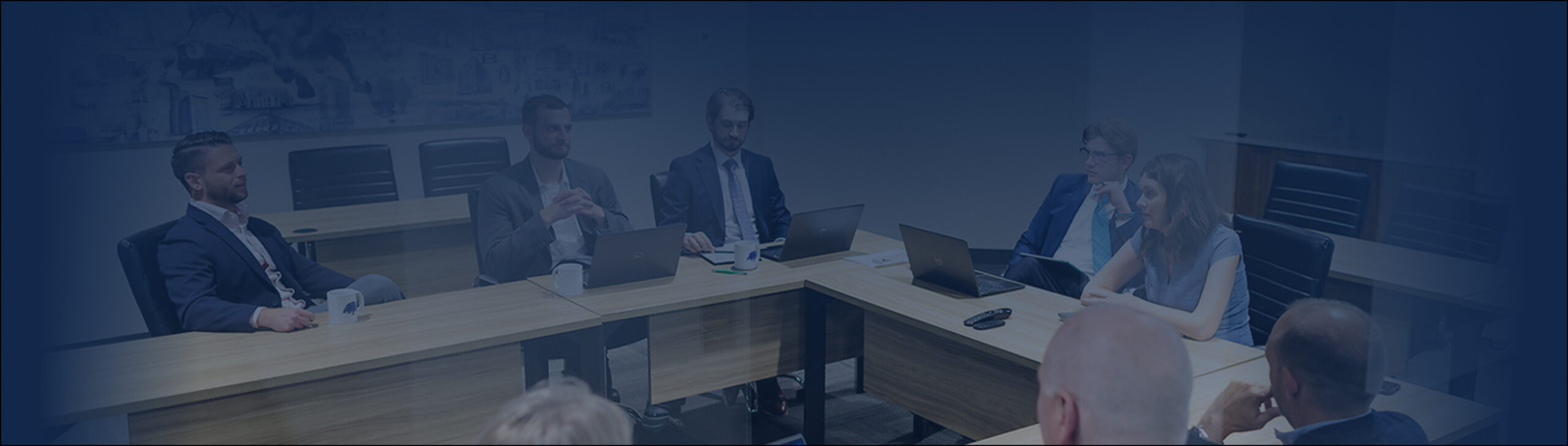 A group of business professionals sit around a U-shaped conference table in a modern meeting room, some with laptops and notebooks, engaged in collaborative discussion.