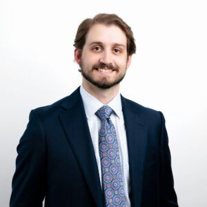 Tyler Johnson, a man with light brown hair and a beard, is wearing a dark blue suit, patterned tie, and checked shirt, standing against a plain white background and smiling at the camera.