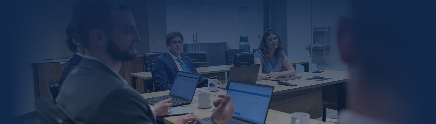 A group of people in business attire sit around a conference table with laptops, engaged in a meeting focused on financial planning and investment in a modern office setting.