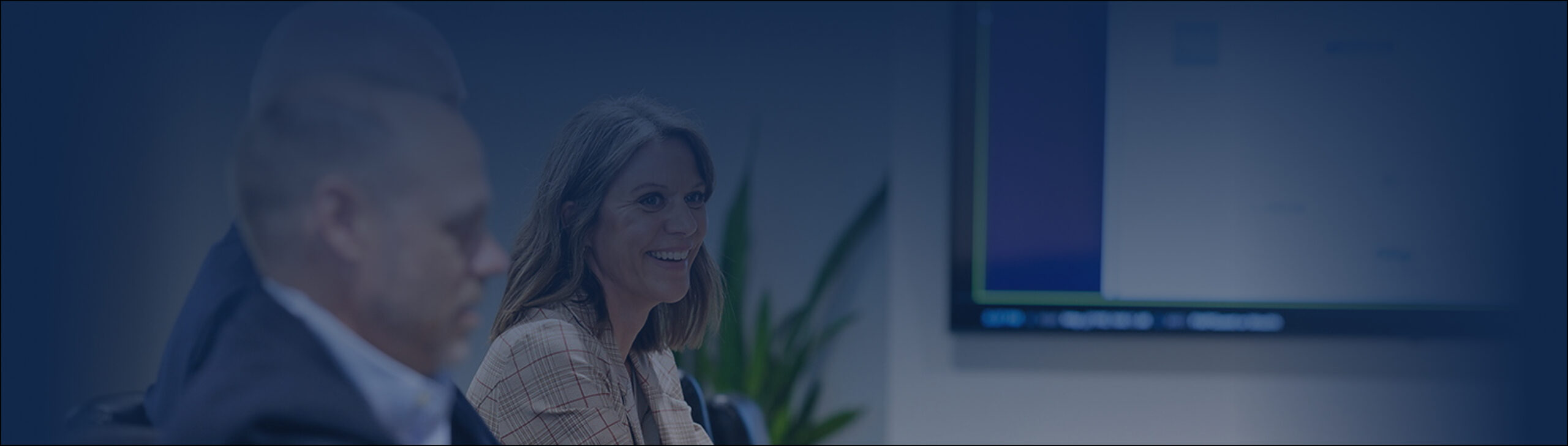 A woman smiling while sitting at a conference table with two men; a blurred screen and plant are visible, reflecting why clients choose us.