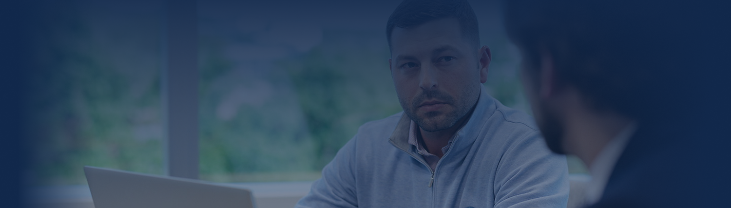 Two advisors sit at a table in an office setting, one facing the camera with a neutral expression and a laptop in front of him, while the other is partially seen from behind. The background is blurred.