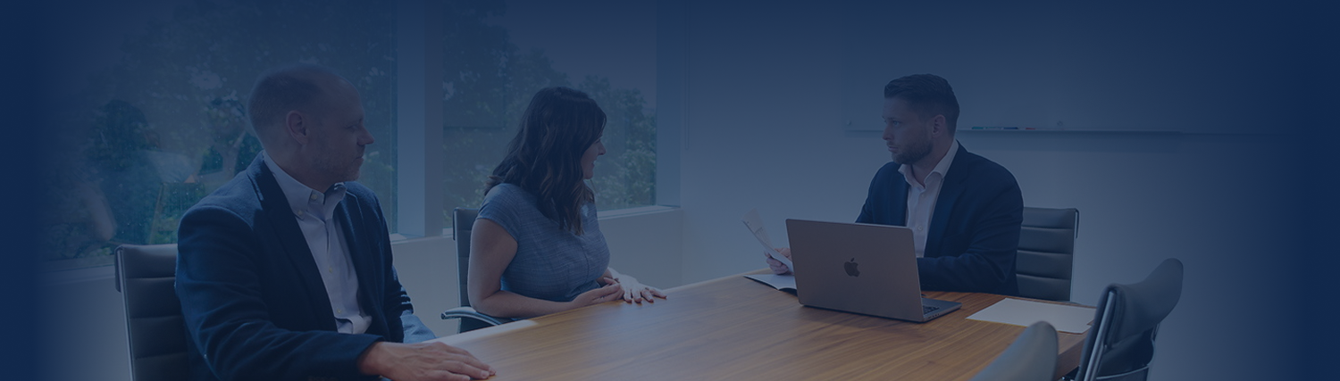 Three people in business attire sit around a conference table in a modern office, engaged in a meeting. One person uses a laptop while talking to the others. Large windows showcase trees outside, creating a collaborative atmosphere.