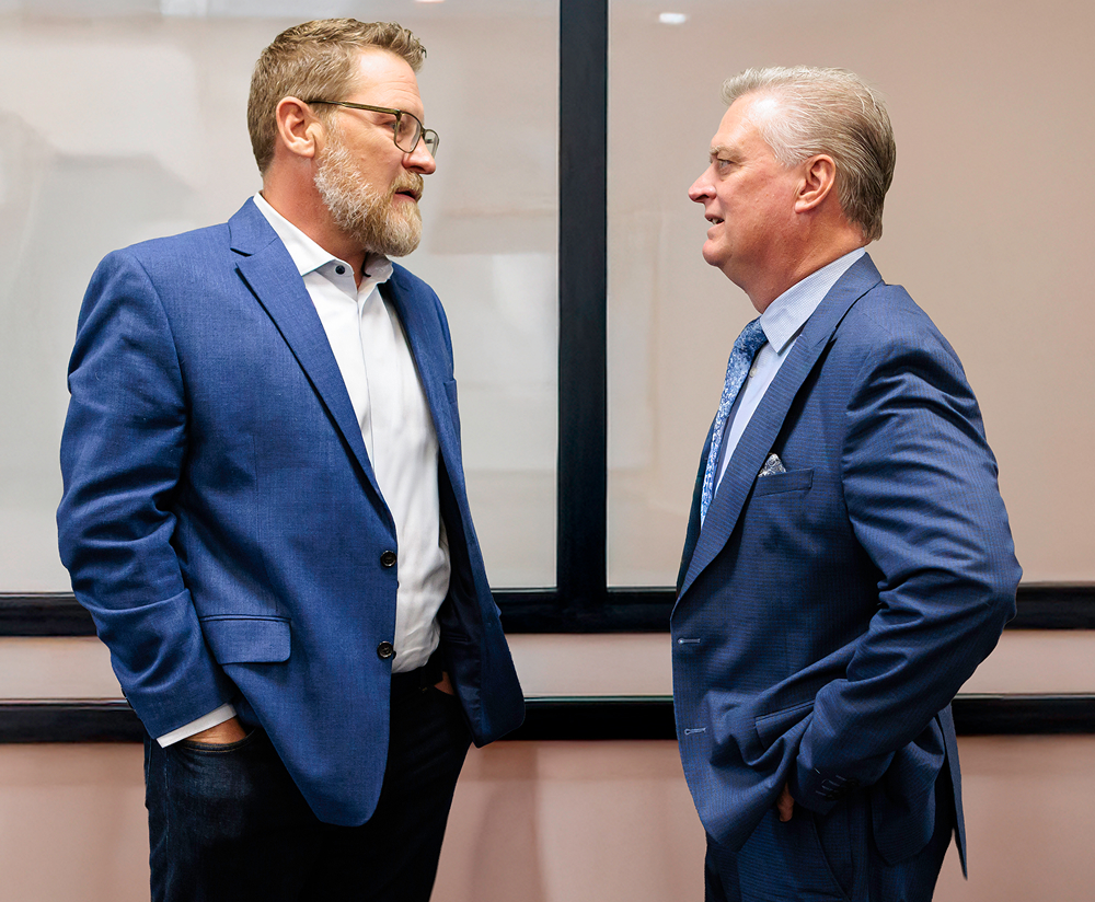 Two men in blue suits stand indoors, engaged in a conversation about financial planning. One has a beard and glasses, the other gray hair. Both have their hands in their pockets as they discuss important matters.