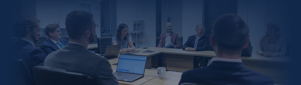 A group of eight people in business attire sit around a conference table, engaged in tax planning discussions, with laptops and papers in front of them in a modern office meeting room.