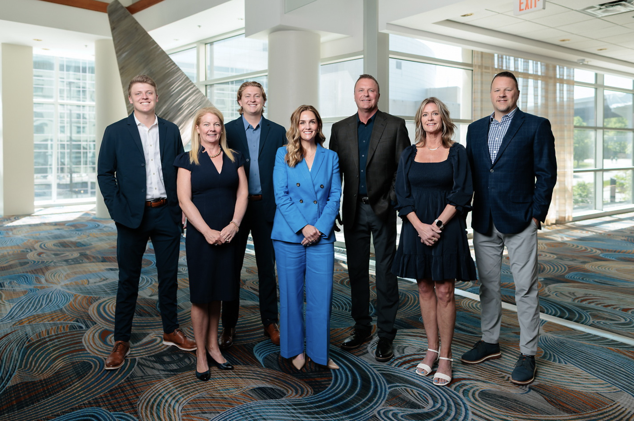 Seven people, dressed in business and business-casual attire, stand together indoors on a patterned carpet in a brightly lit, modern space with large windows—perhaps gathered to discuss the company’s new retirement plan.